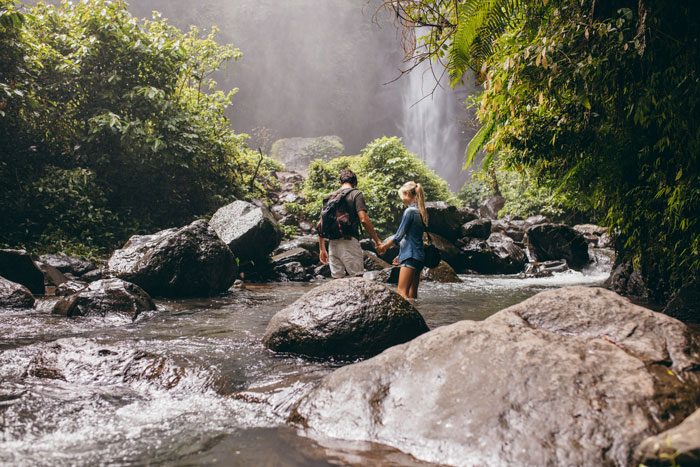 Mladi par se drži za ruke dok stoje u potoku sa slapom u pozadini